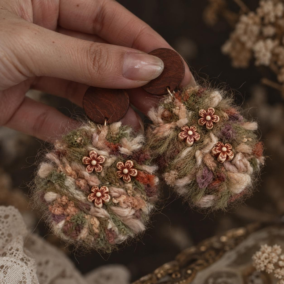 Front of the cream and green yarn floral earrings with brown studs, held in a hand.